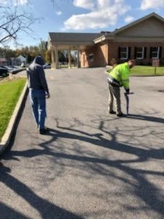 Two technicians in high-visibility gear locating utilities in a parking lot near a building.