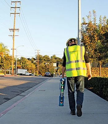 Technician in a high-visibility vest walking along a sidewalk while carrying a Radiodetection utility locator.