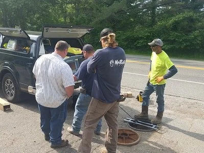 Crew gathered around an open manhole using inspection equipment near a vehicle.