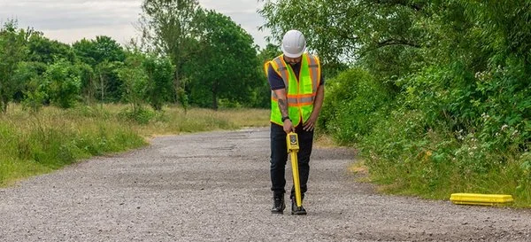 Technician in a hard hat and safety vest using a Radiodetection locator on a gravel path surrounded by trees.