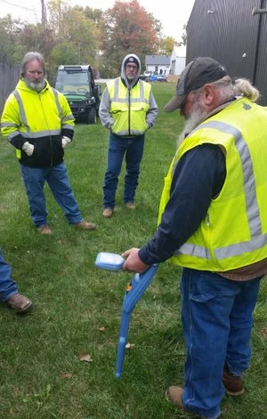 Technician in a high-visibility vest demonstrating a utility locator wand on grass while others observe.