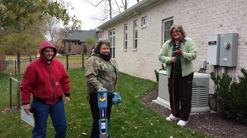 Three people talking outdoors in a residential side yard near a utility unit.
