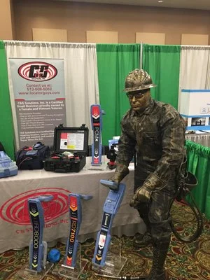 C&S Solutions trade show booth with a green curtain backdrop and a person in camouflage attire.