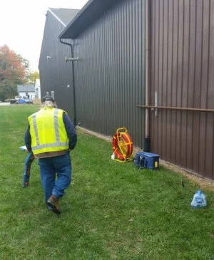 Technician in a high-visibility vest walking beside a barn with locating equipment set on the grass.