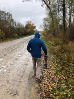 Technician walking alone along a gravel road through autumn trees.