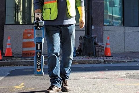Technician carrying a Radiodetection RD8200 locator on a city street near traffic cones and a utility marking.