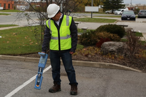 Technician in hard hat and safety vest holding a utility locator transmitter while surveying a parking lot.