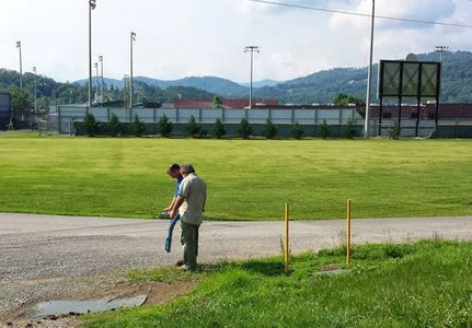 Technician using a locator on a paved walkway near a sports field.