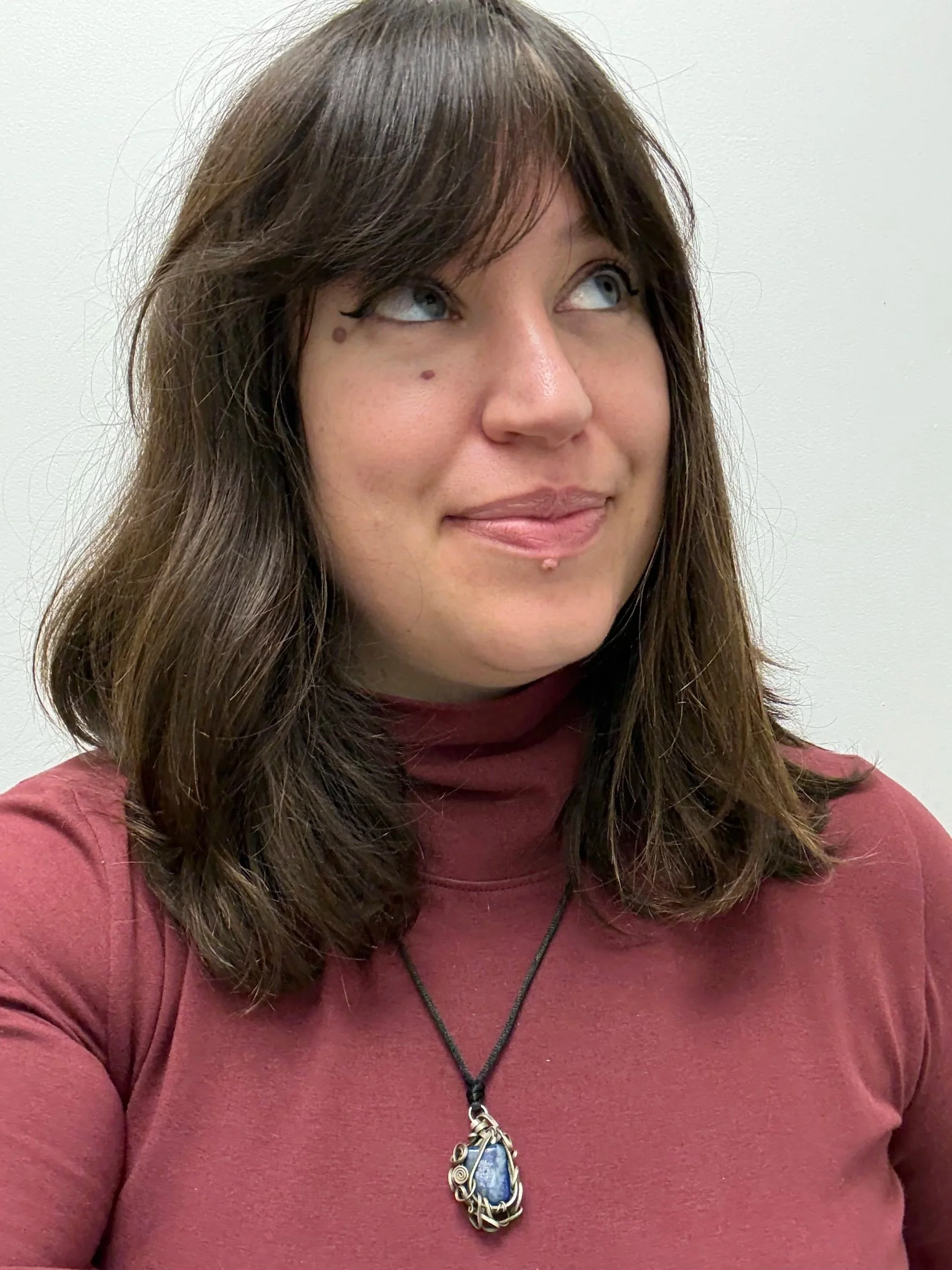 A woman with shoulder-length brown hair, wearing a maroon long-sleeve shirt and a large blue pendant necklace, standing against a plain white wall.