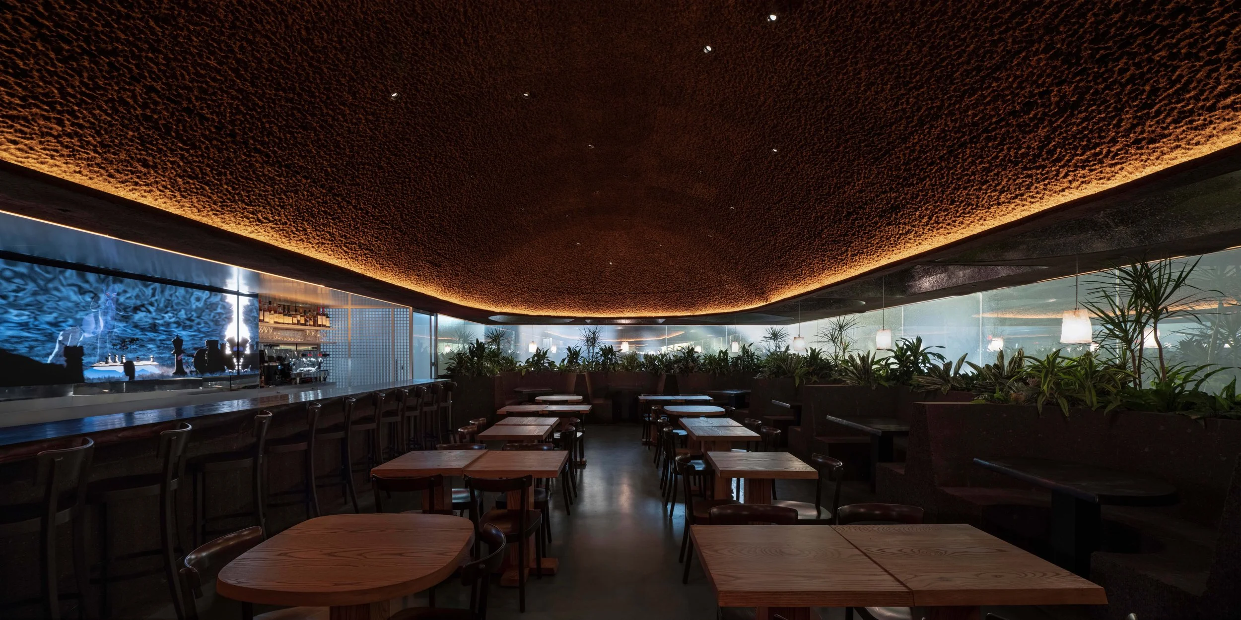 Interior of a modern restaurant with wooden tables and chairs by Minjae Kim, lush green plants along the window, a textured cork ceiling with integrated lighting, and a bar area visible in the background with bar art by Ian Cheng.