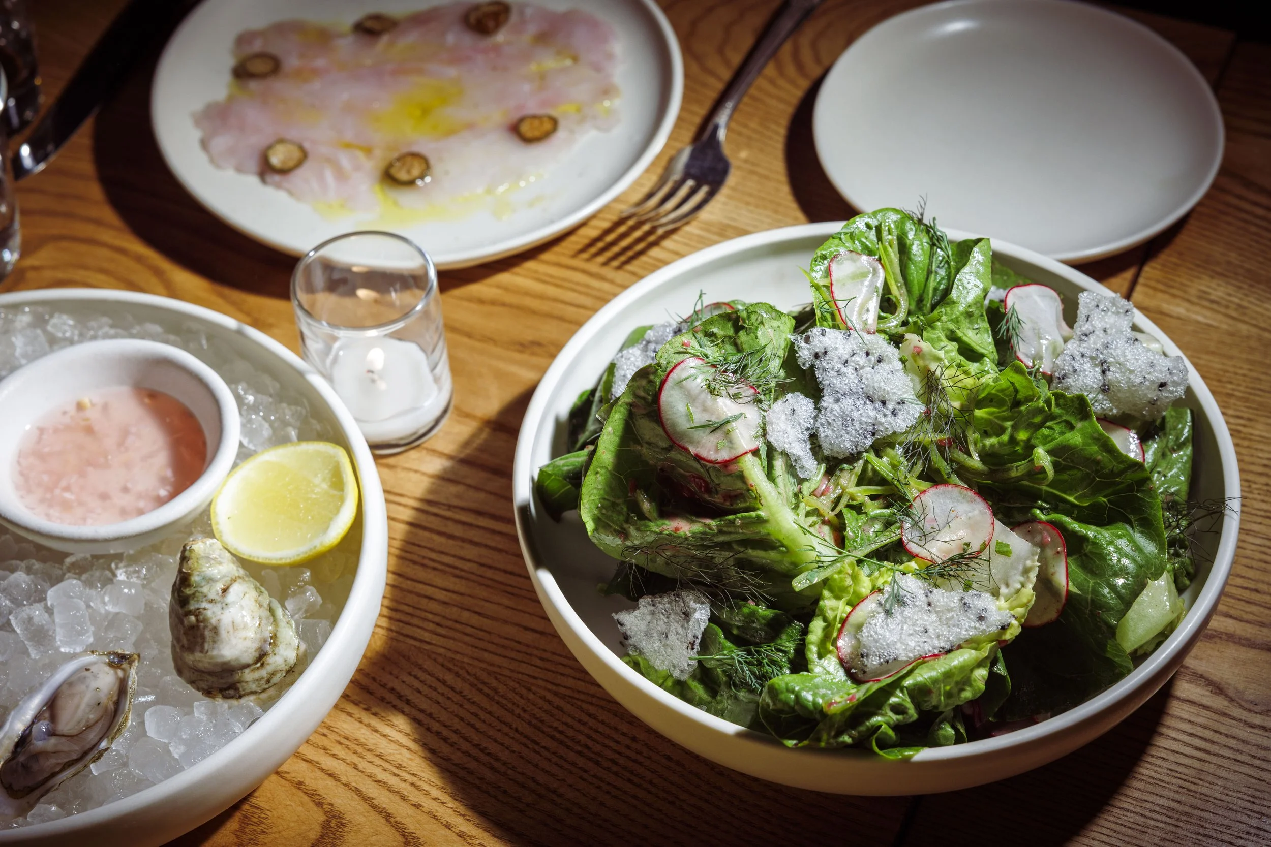 A table with a bowl of Little Gem salad topped with shaved radishes, herbs, and crispy cheese, a plate of crudo with olives and olive oil, and oysters. 