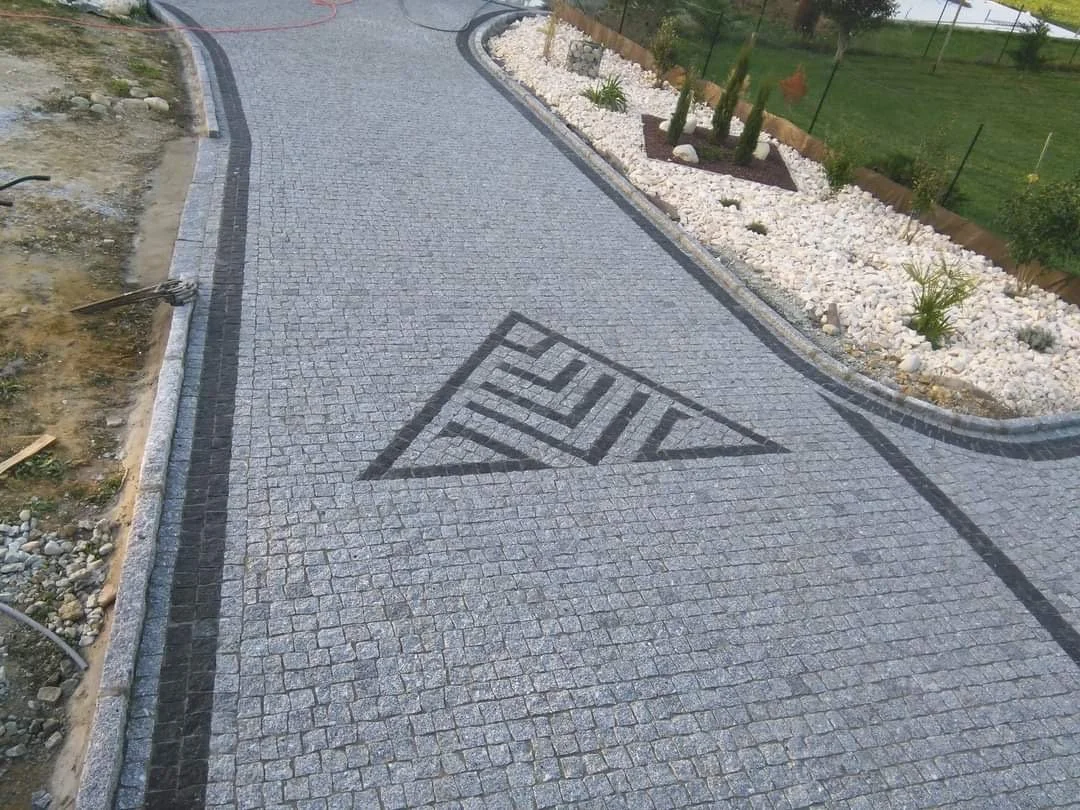 Une allée pavée avec un motif en forme de triangle noir et blanc, bordée de pierres et de plantes, dans un jardin