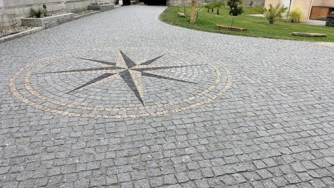 Une allée pavée en pierre avec un motif de rose des vents au centre, entourée d'un jardin avec des arbres et des plantes.