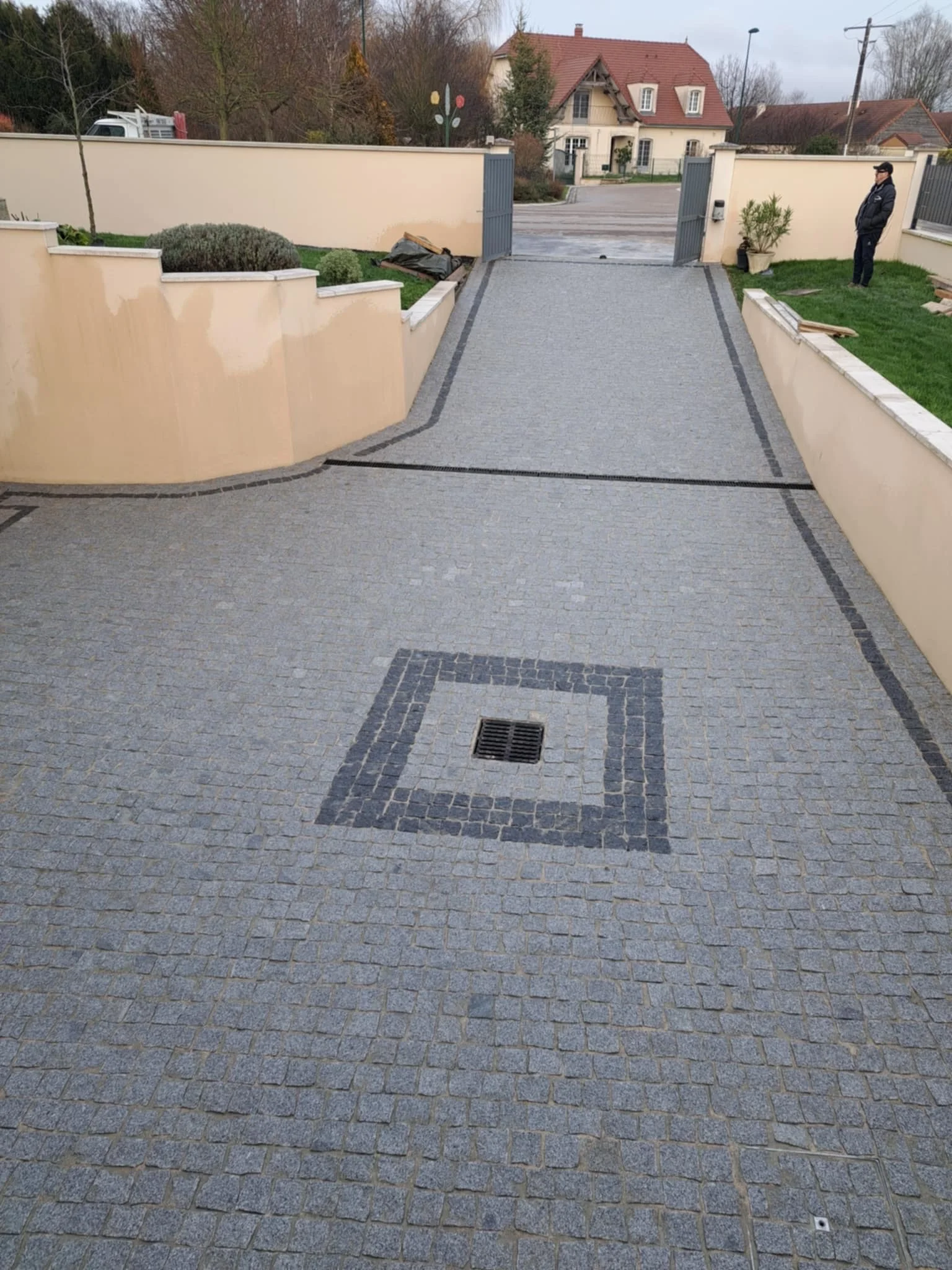 Une entrée de maison avec un portail ouvert, une allée pavée en pierre, un mur en béton beige, un homme en veste noire sur la droite, et des maisons résidentielles en arrière-plan.