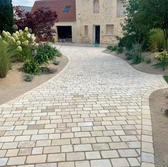 Allée pavée sinueuse entourée de jardin avec fleurs et arbustes, menant à une maison en pierre.
