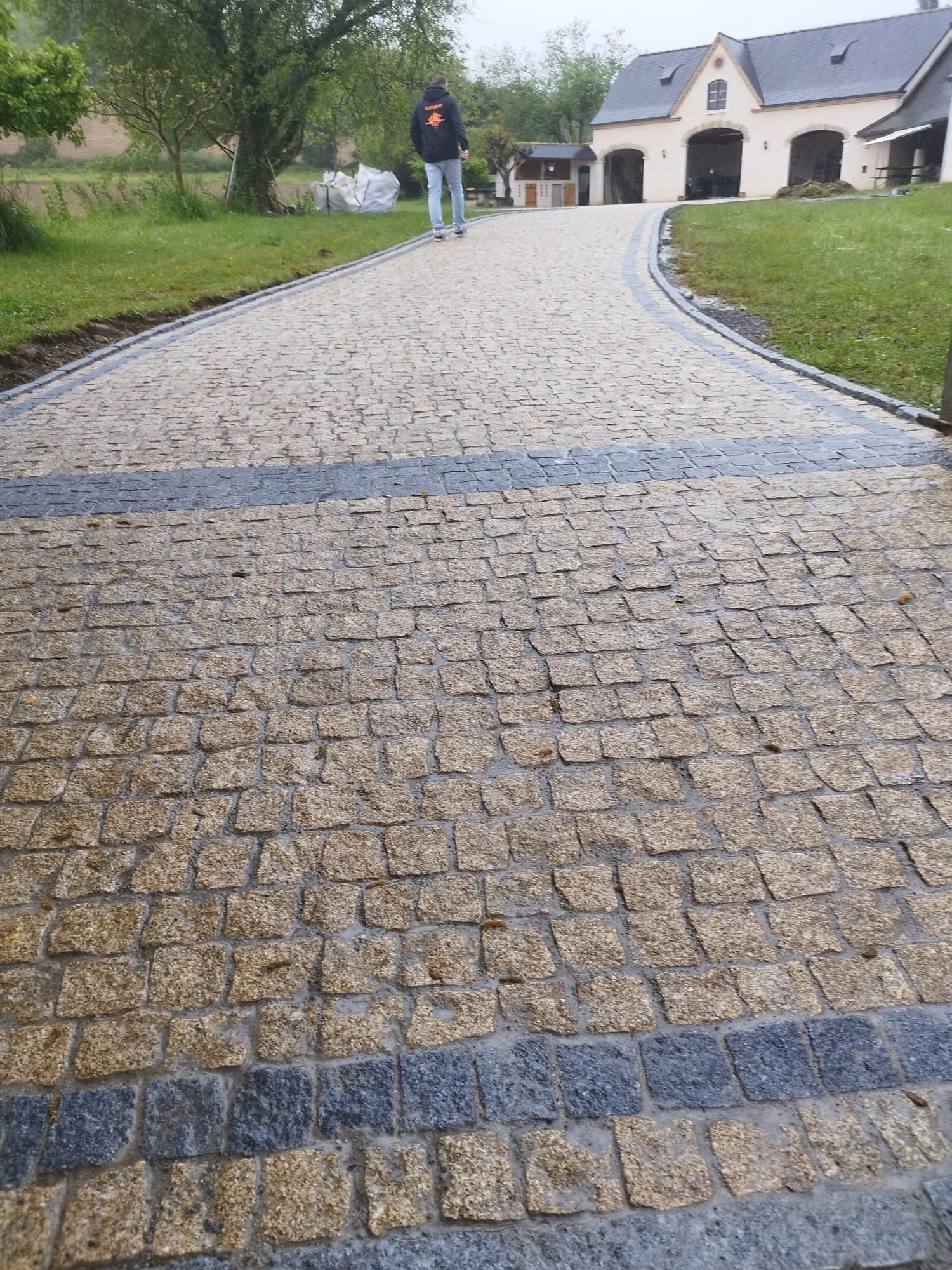 Une allée pavée menant à une maison, avec un homme qui marche, entourée d'arbres et de pelouses verdoyantes.