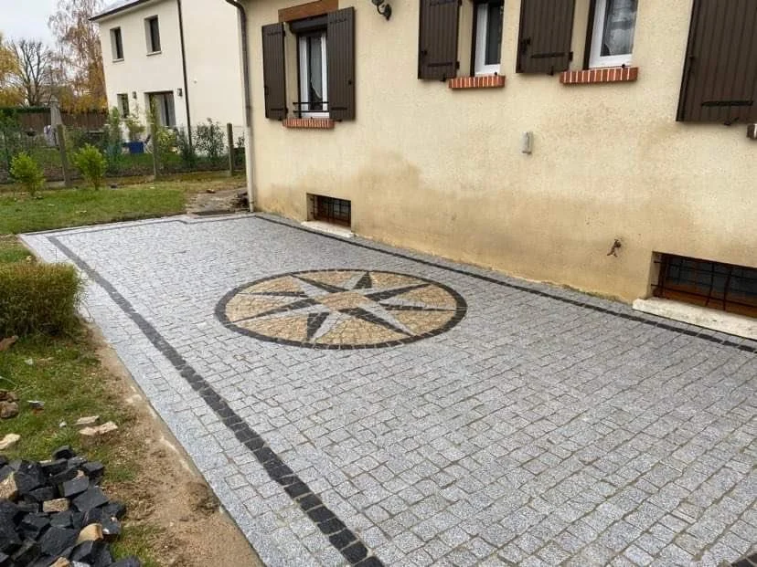 Cour pavée avec un motif circulaire au centre, adjacent à une maison avec volets en bois.