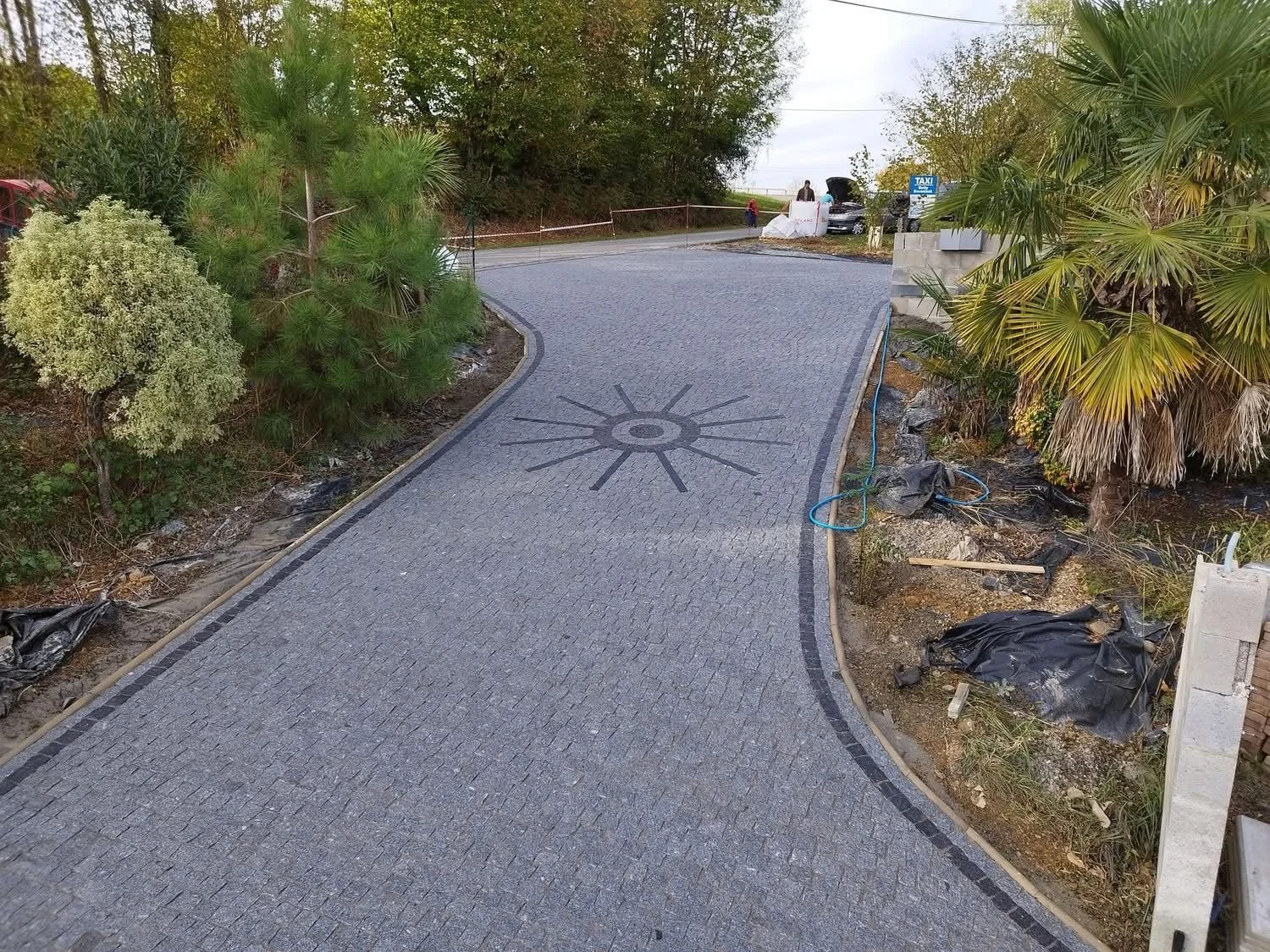 Un chemin de pavés en construction avec un motif circulaire en centre, entouré de végétation, avec des travaux en cours sur le côté droit.