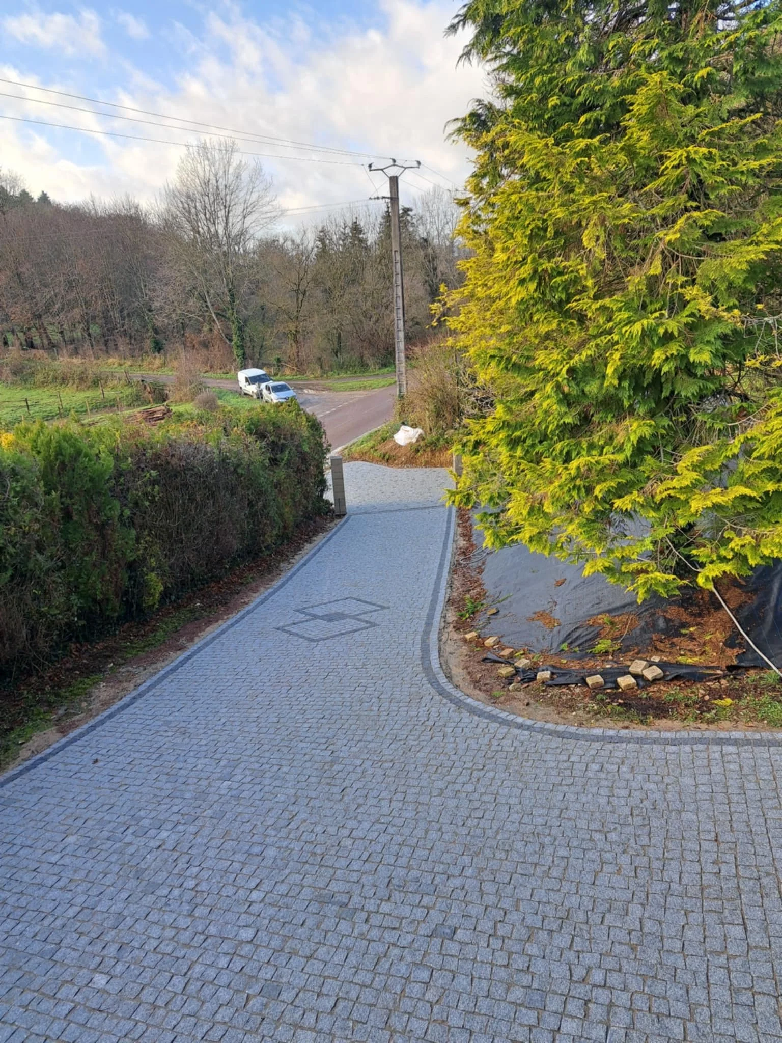 Un chemin pavé serpentant à travers un paysage rural avec des arbres verts et nus, une voiture blanche et une voiture grise stationnées au croisement, un poteau électrique, et un ciel partiellement nuageux.
