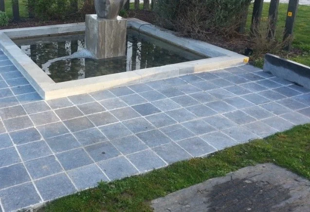 Une fontaine avec une structure en pierre dans une fontaine d'eau entourée de pavés gris, située sur une pelouse.
