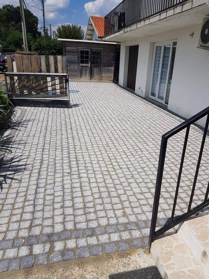 Une terrasse pavée devant une maison avec une porte-fenêtre, un petit cabanon en bois à l'arrière, et une partie de l'escalier en métal à droite.