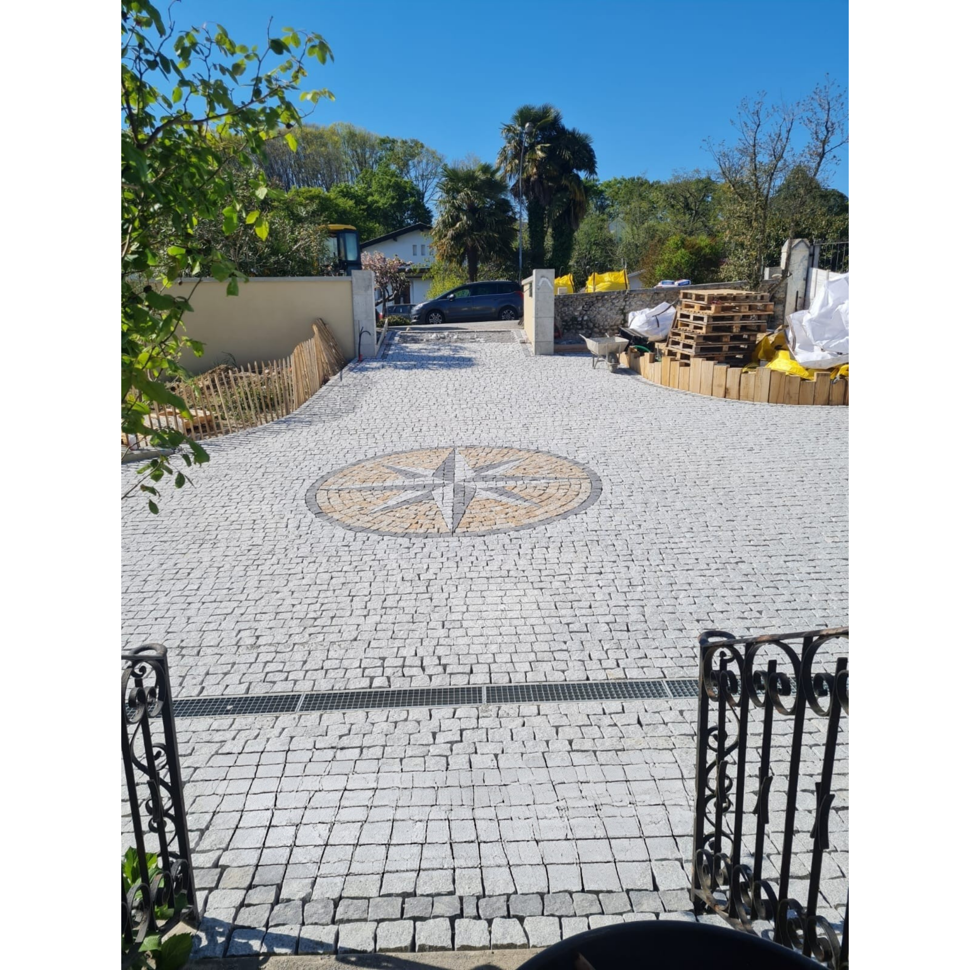 Un chemin pavé menant à une entrée de maison entourée de murs et de végétation, avec des arbres, des palettes en bois, et des matériaux de construction sur le côté droit, sous un ciel bleu clair.
