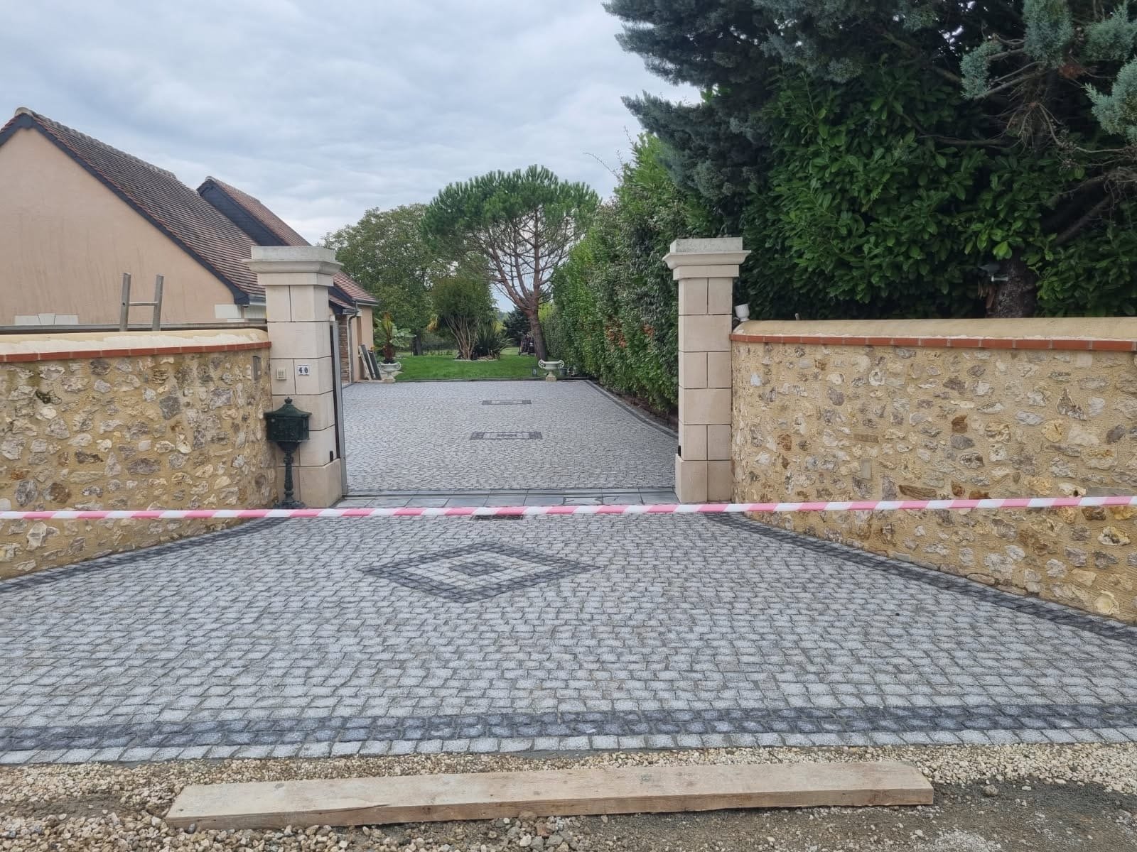 Entrée d'une propriété avec un portail ouvert, un chemin en pavés, un mur en pierre de chaque côté, et un jardin avec des arbres et de la pelouse en arrière-plan.