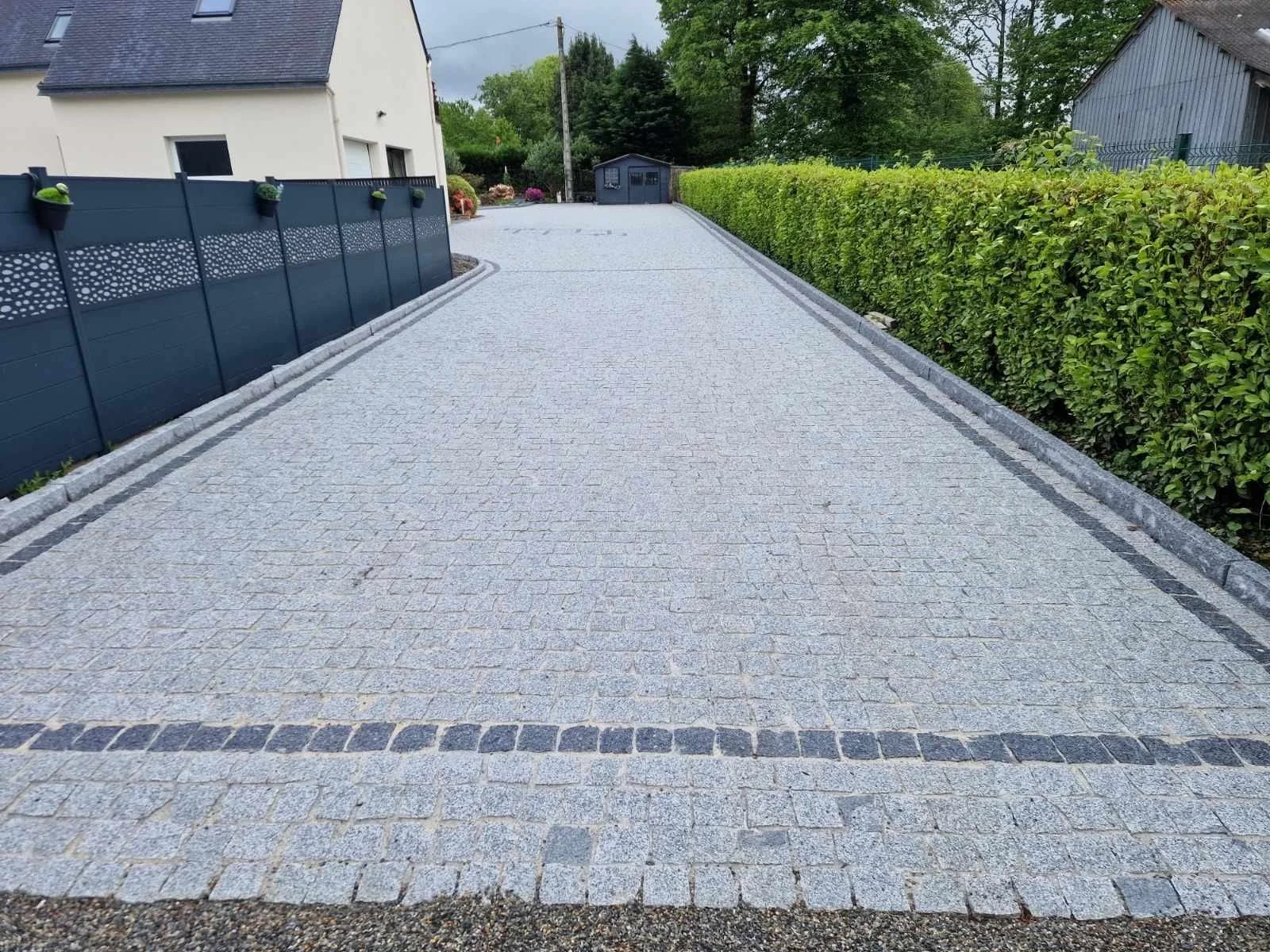 Chemin pavé en pierre avec bordure en pierres de couleur plus foncée, flanqué d'un pavage de haie verte dense à droite et d'une clôture noire décorative à gauche, avec une maison blanche visible en arrière-plan.