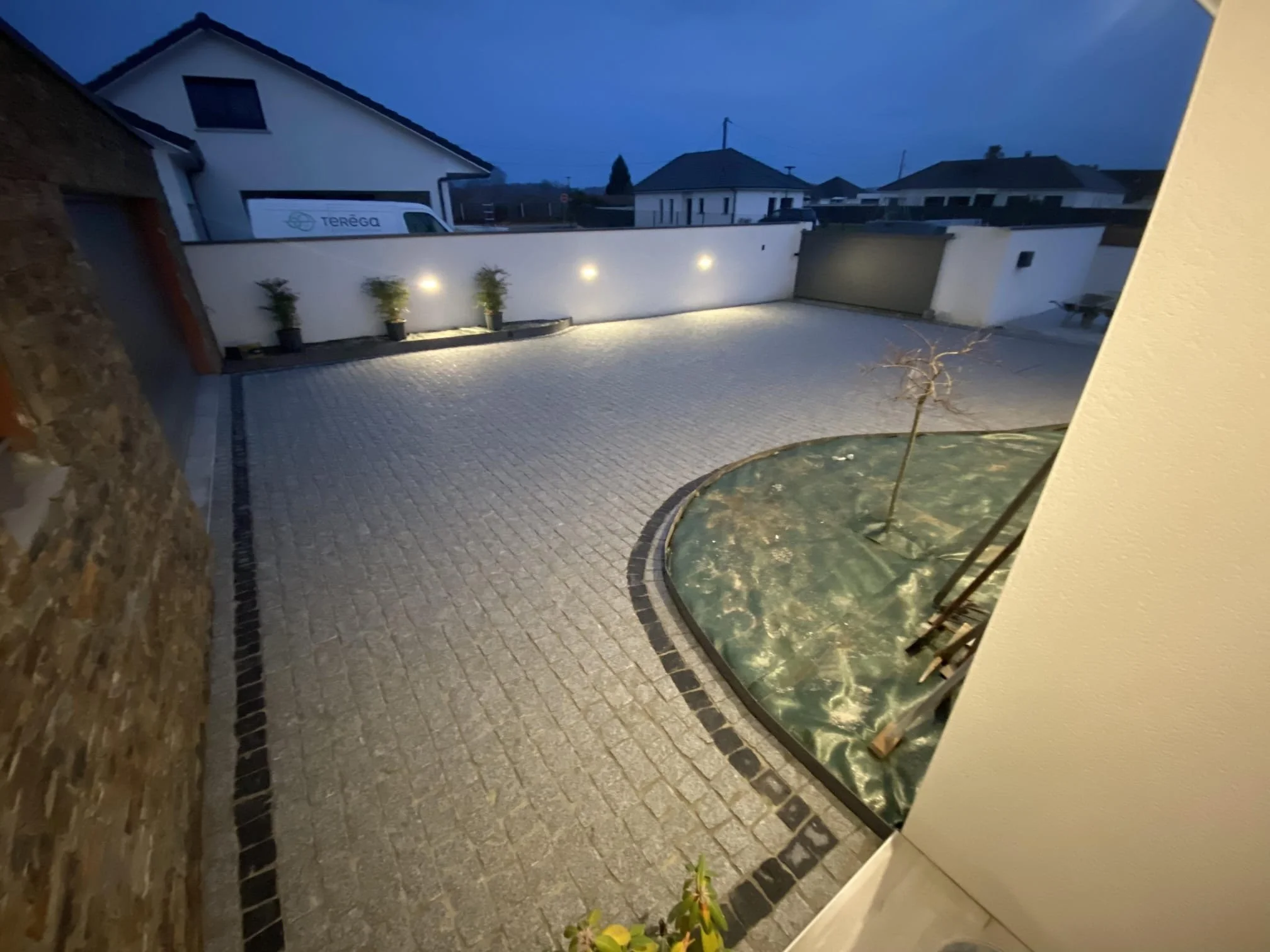 Un patio éclairé la nuit avec un mur blanc, deux plantes en pot, un arbre petit dans une zone de gravier, des maisons environnantes et un ciel sombre.