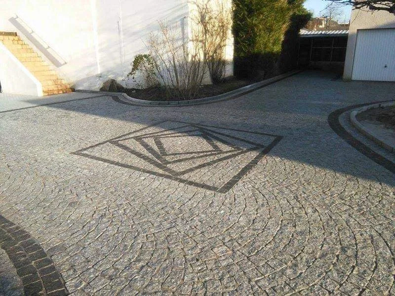 Une cour en pavés avec un dessin géométrique en pierre sur le sol, des escaliers en face, une maison avec un garage, et des arbres en arrière-plan.