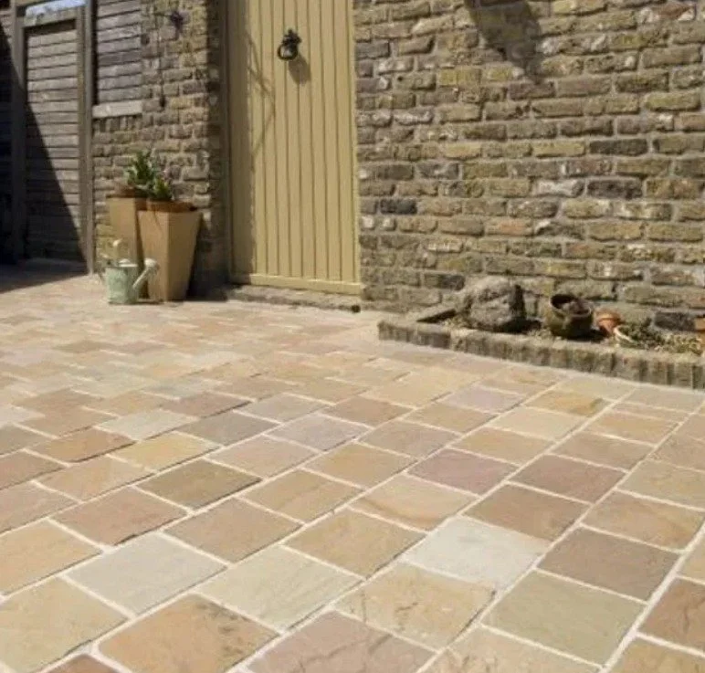 Cour en pierre avec une porte en bois beige, un mur en briques, et des pots de fleurs près de la porte.