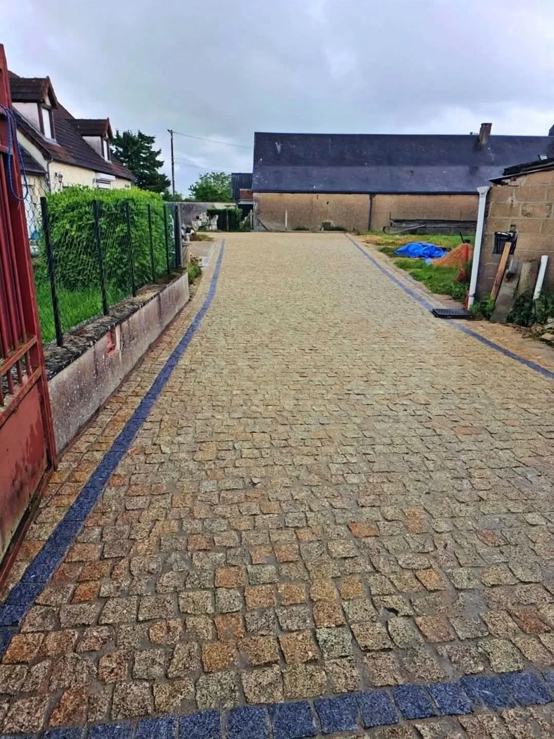 Chemin en pavés avec bordures bleues, côté gauche clôture en métal vert et côté droit bâtiment en parpaings.