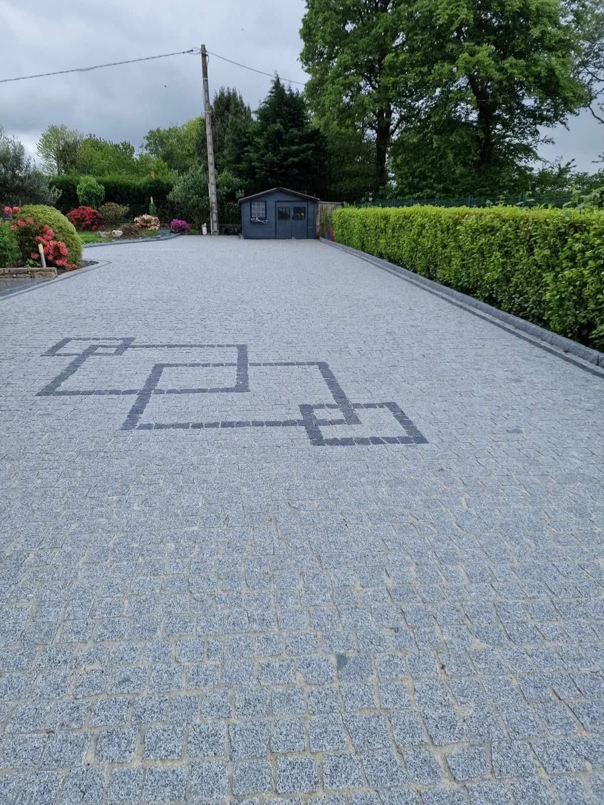 Une allée pavée avec une fresque de deux cases à jouer au jeu de dames en pierre, entourée de haies vertes et de plantes fleuries, avec un petit bâtiment en arrière-plan et un ciel nuageux.