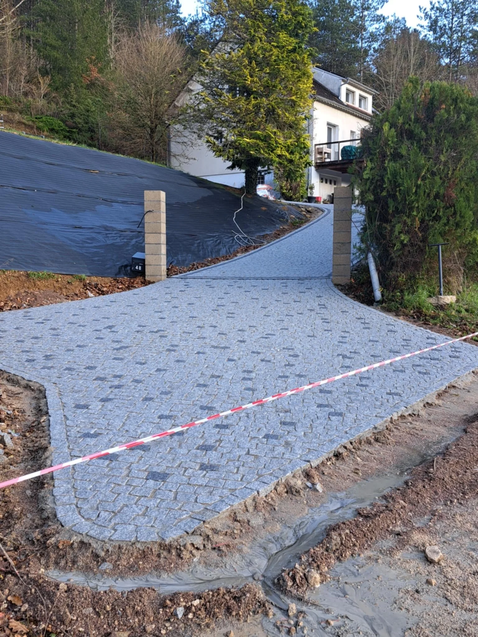 Une allée pavée en pierre en construction devant une maison blanche sur une pente, entourée d'arbres avec de la végétation au printemps.