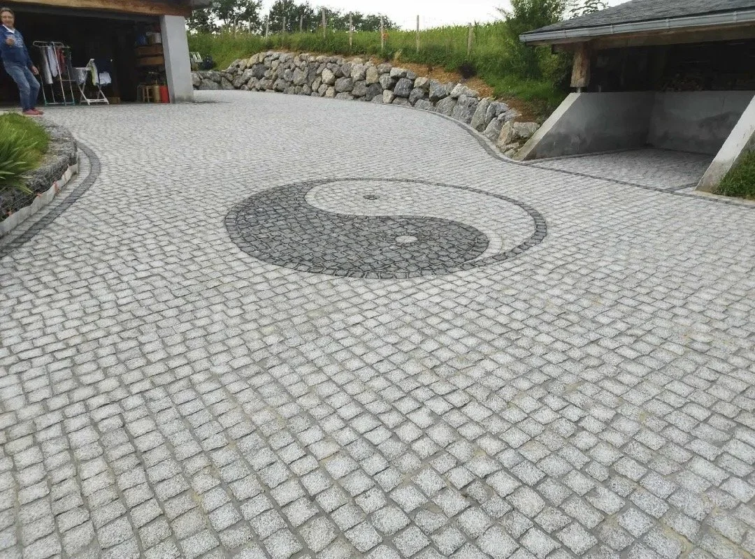 Surface pavée avec un motif yin-yang au centre, entourée de rocailles et de végétation, avec une structure en bois et en béton à l'entrée.