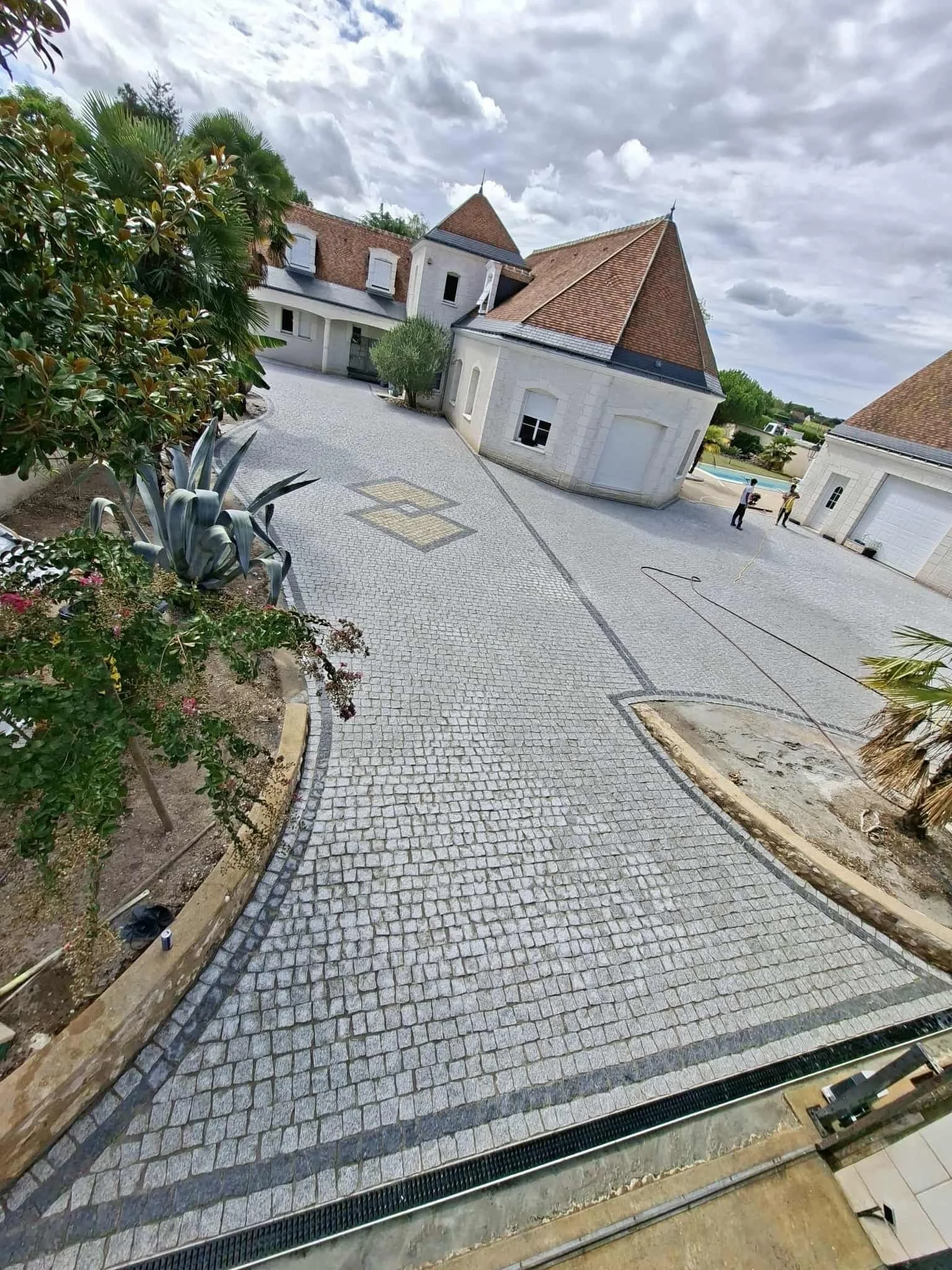 Coulage de pavés en cours dans une cour, avec des travailleurs. Bordures de pavés visibles et végétation environnante.