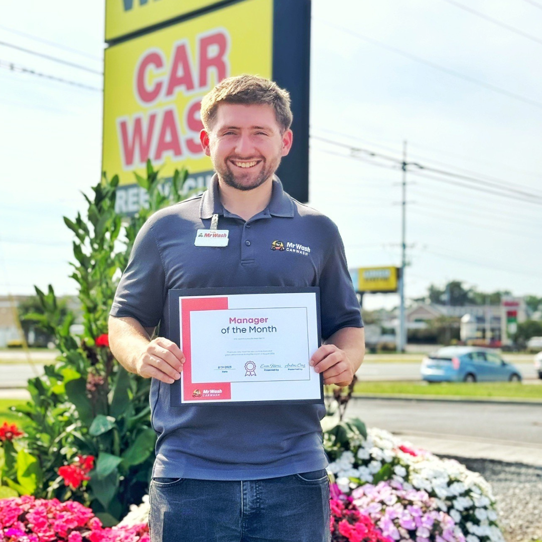A man smiling and holding a certificate that says "Manager of the Month" outdoors near a flower bed, with a yellow and red sign for Mr. Wash Car Wash in the background.