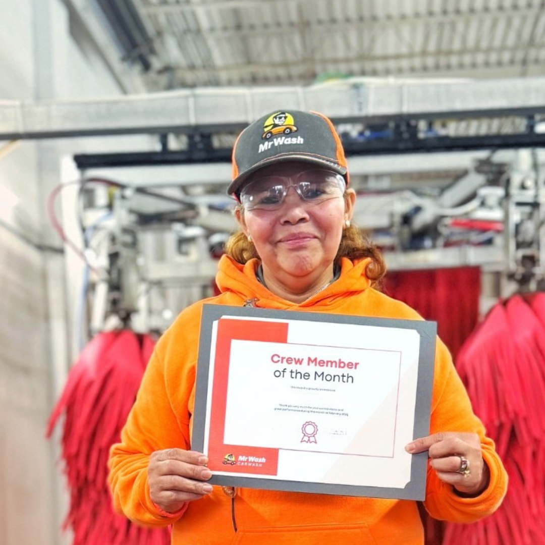 A woman wearing safety glasses, a black and orange cap with a logo, and an orange hoodie, holding a certificate that reads 'Crew Member of the Month', in a laundry or industrial setting with red cleaning cloths hanging behind her.