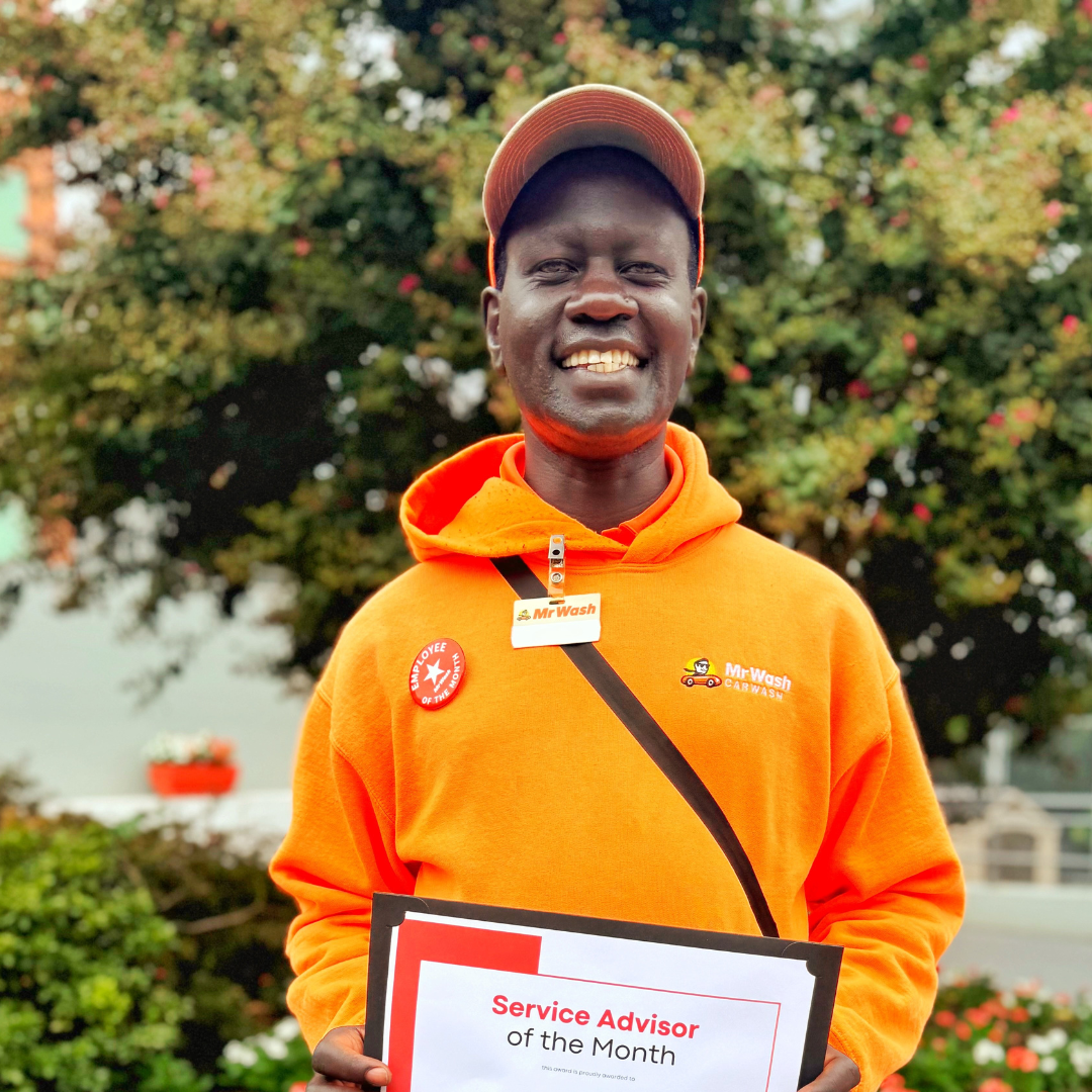 A smiling man wearing an orange hoodie, cap, and a badge, holding a certificate for Service Advisor of the Month outdoors with trees in the background.