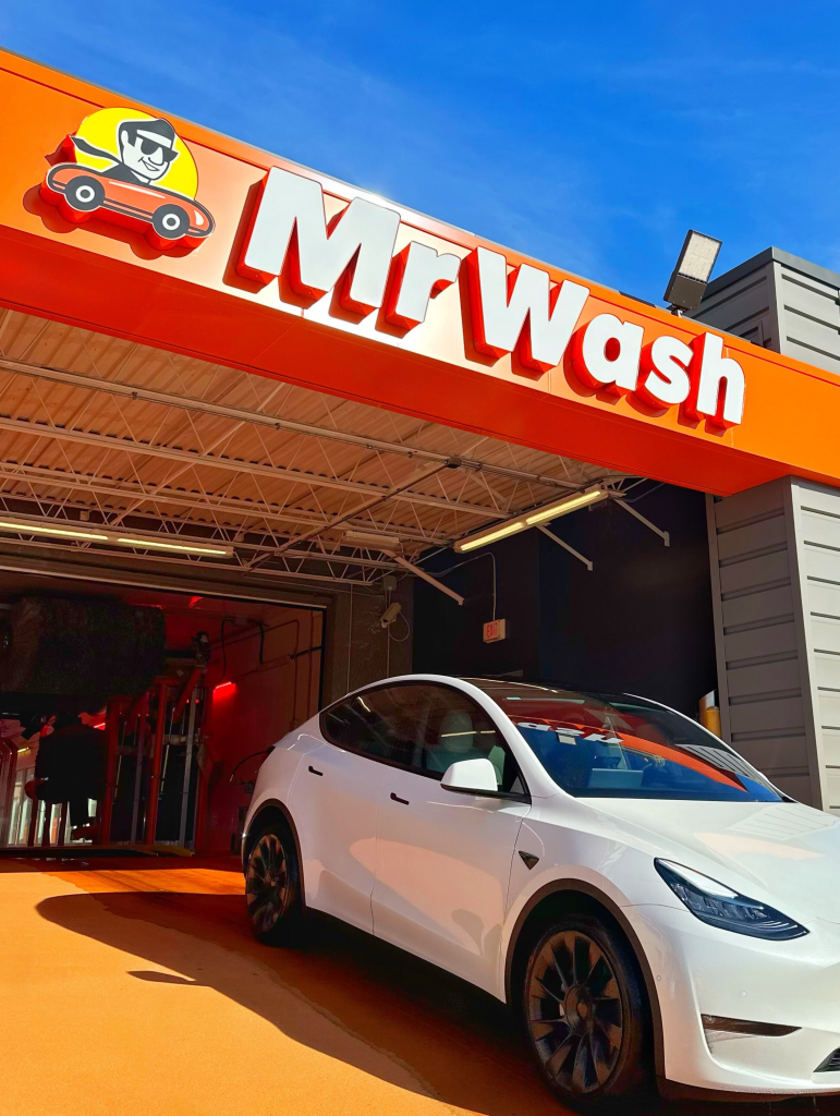 A Tesla Model 3 parked outside a Mr. Wash car wash with a bright orange and red sign under a clear blue sky.