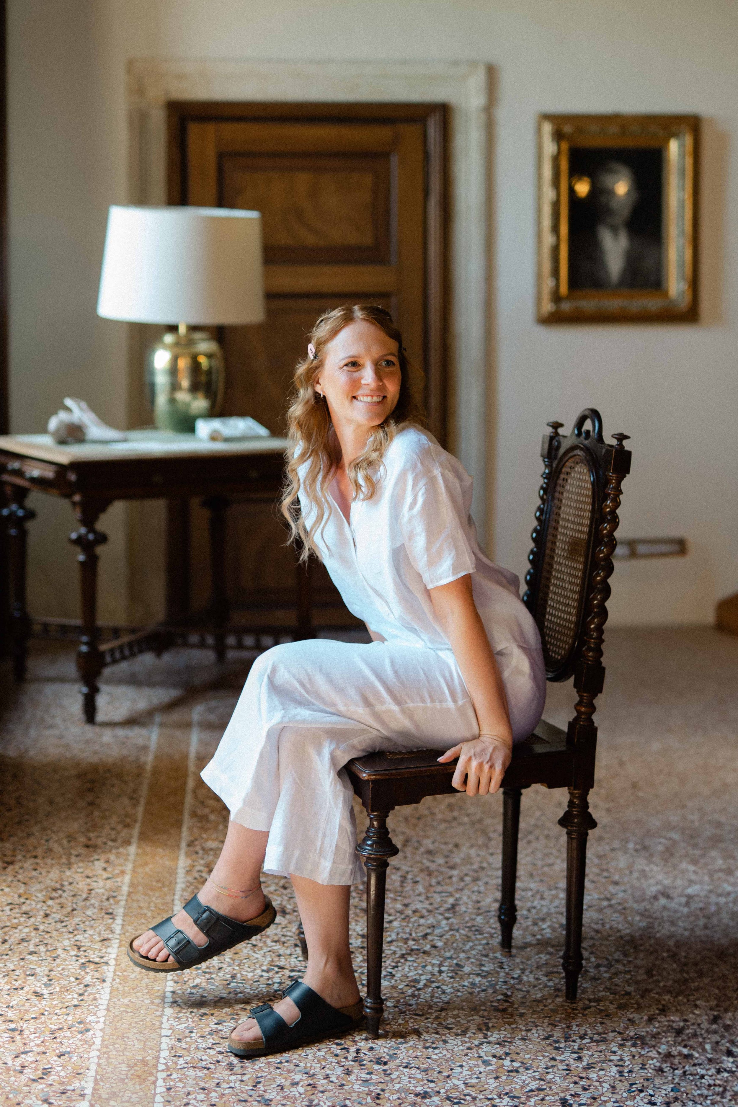 A young woman with long curly blonde hair, wearing a white dress and sandals, sitting on a wooden chair in a room with beige walls, a wooden door, a framed painting of a dog, and a table with a lamp.