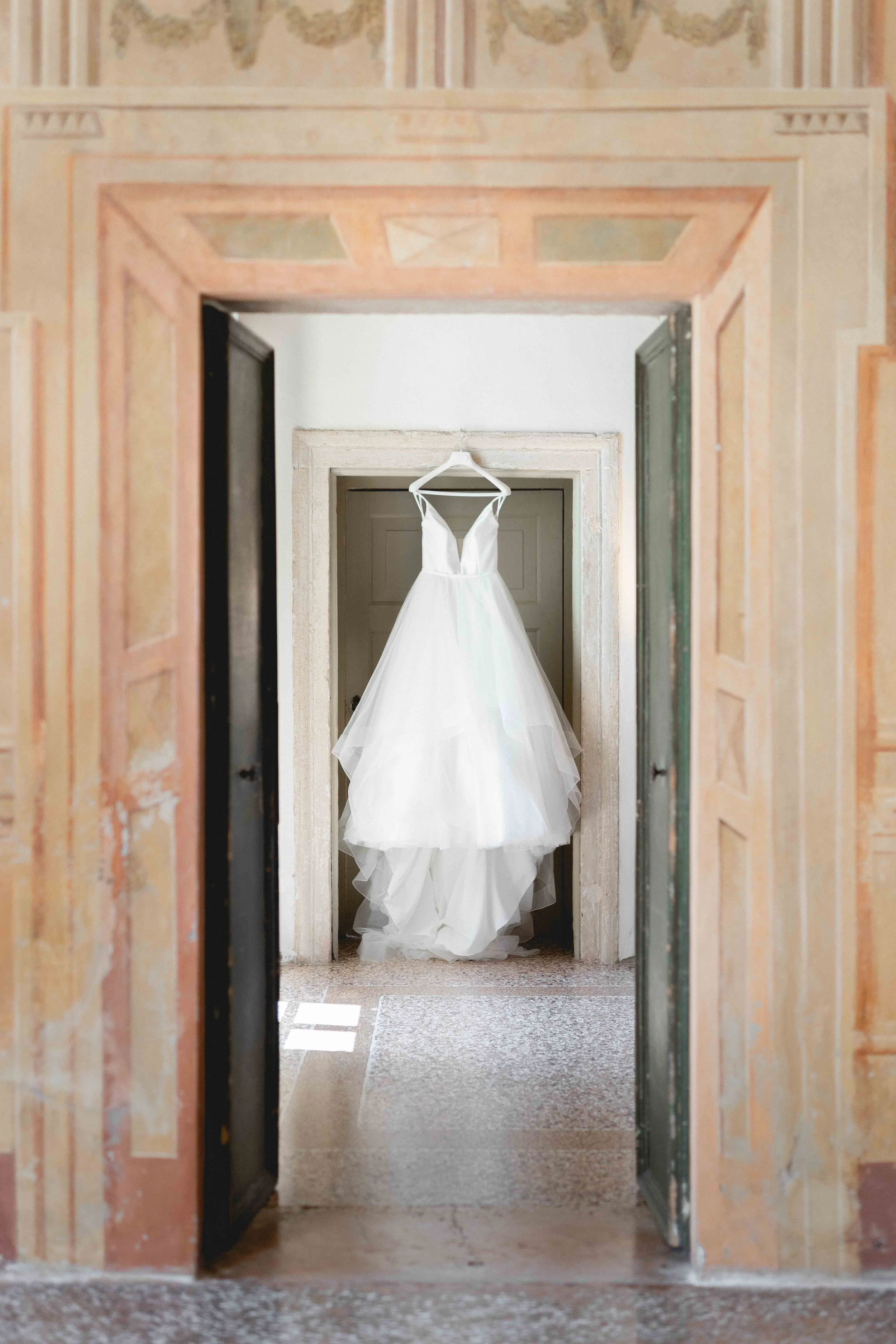 A white wedding dress hanging on a hanger in a doorway, viewed through an open door in an aged building with peach and green painted walls.