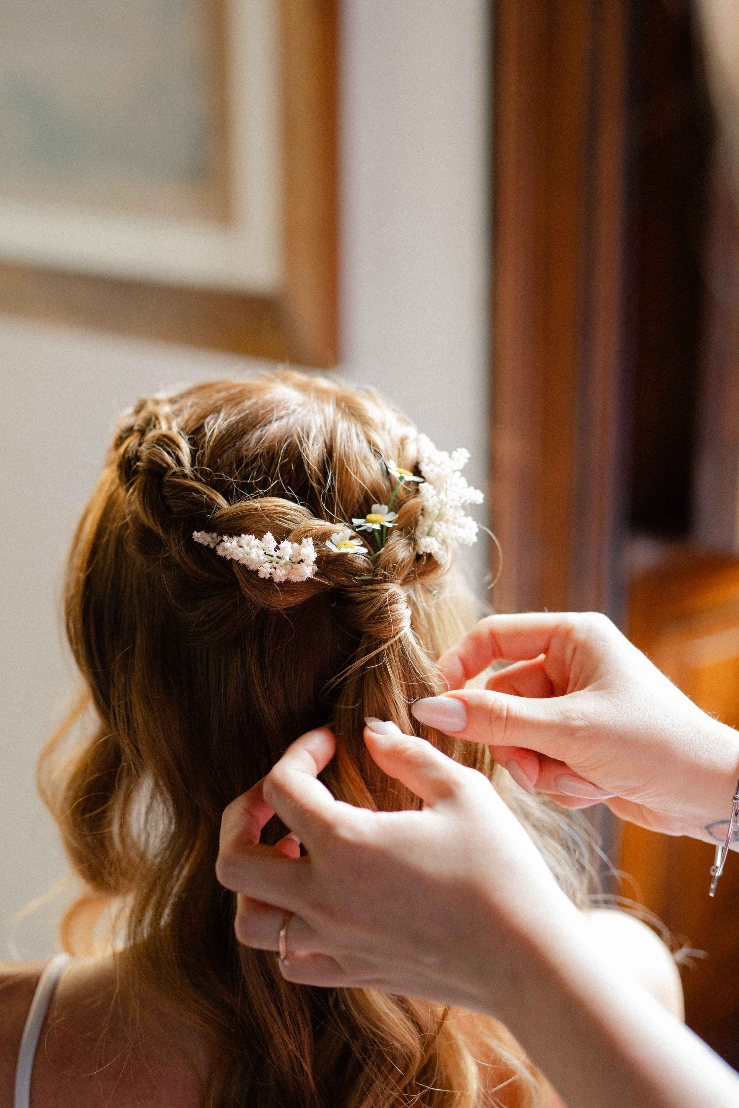 Close-up of a woman with long, wavy, reddish-brown hair, decorated with white and yellow daisies, having her hair styled by a person with light skin, manicured nails, and a gold ring, in a room with wooden furniture.