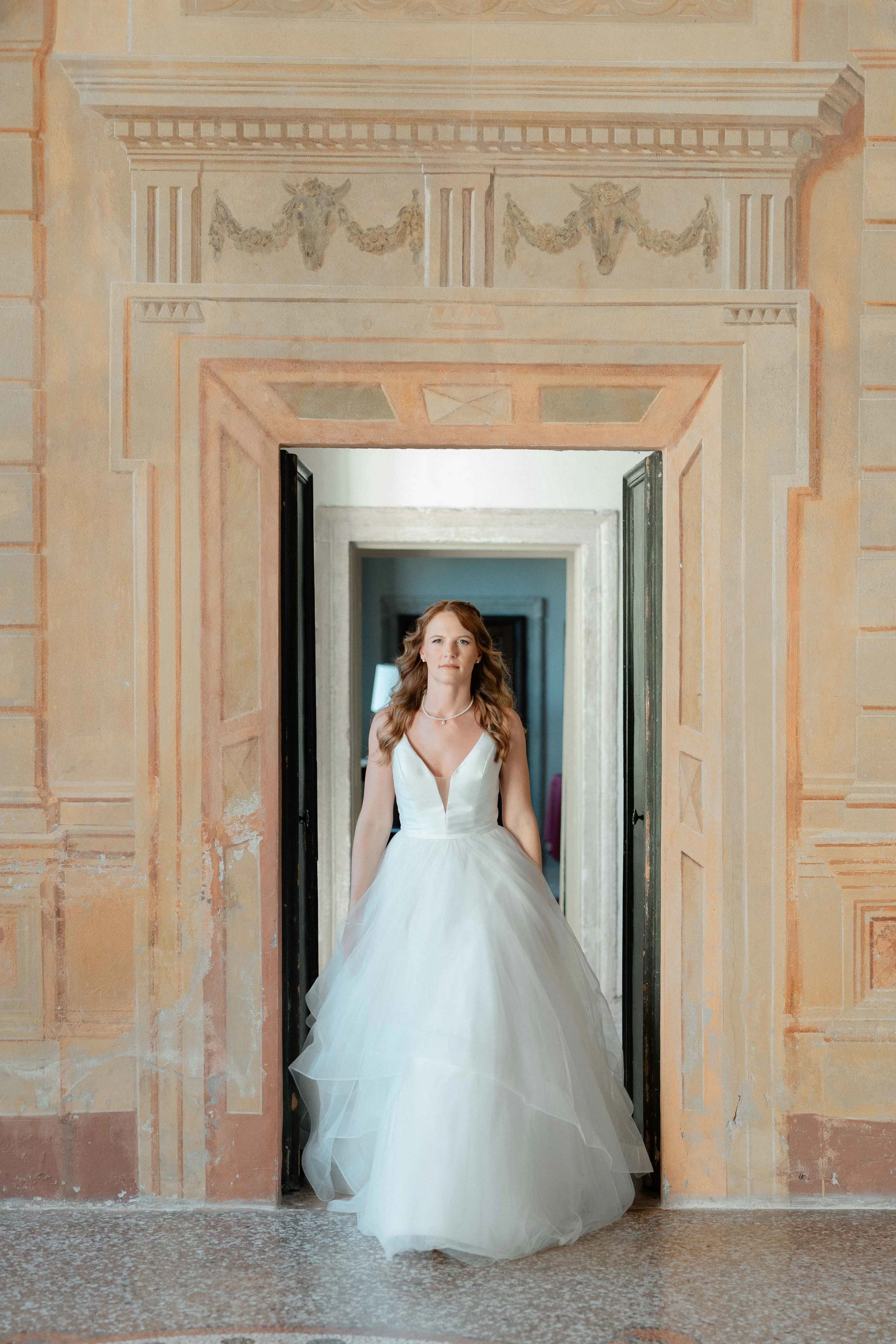 A woman in a white wedding dress standing in a doorway with ornate architecture and painted wall details.