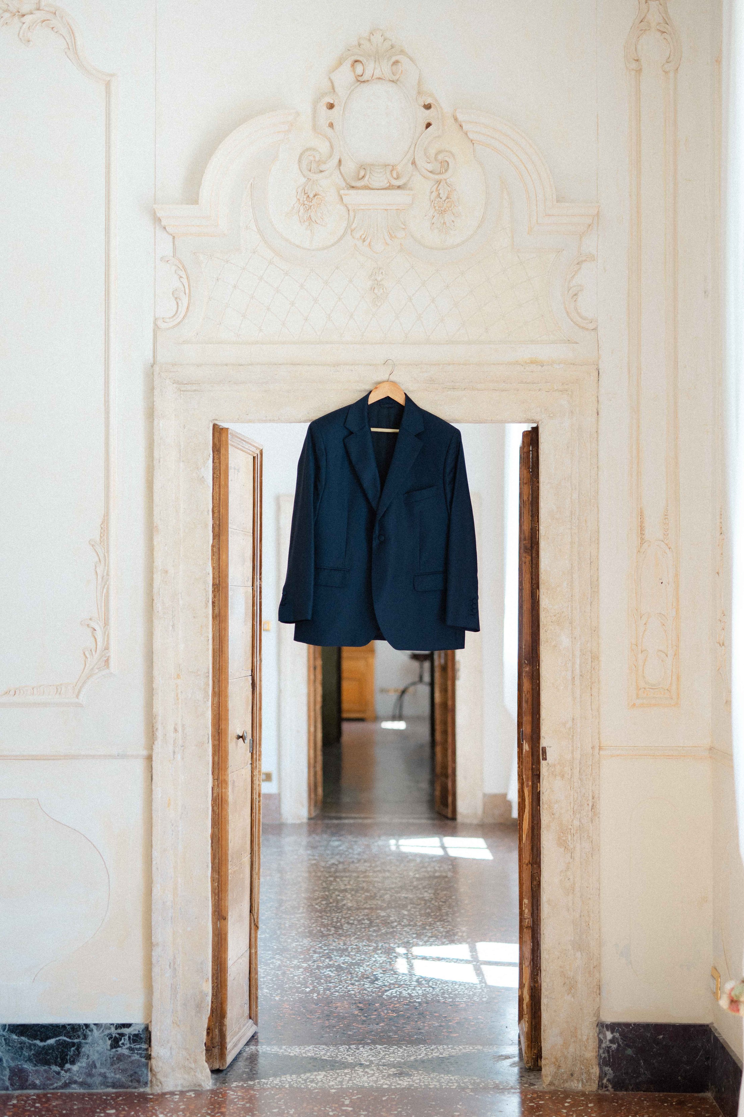 A dark navy blazer jacket hanging on a wooden hanger in a doorway with ornate painted bathroom walls and open double doors leading to a room with a patterned floor.