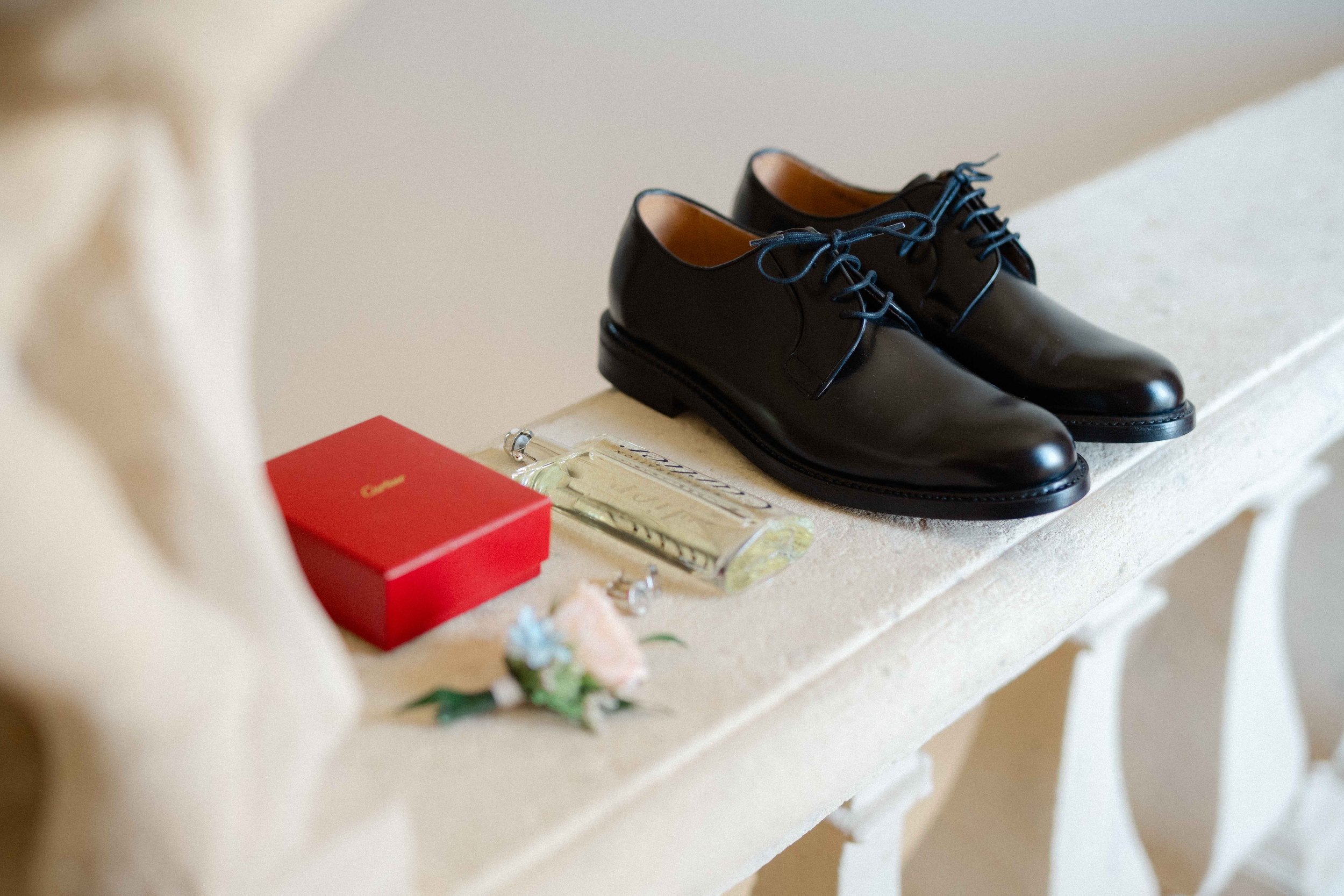 A pair of black dress shoes with blue laces placed on a light-colored surface. Nearby is a red jewelry box, a clear perfume bottle, and some jewelry. The background is blurred.
