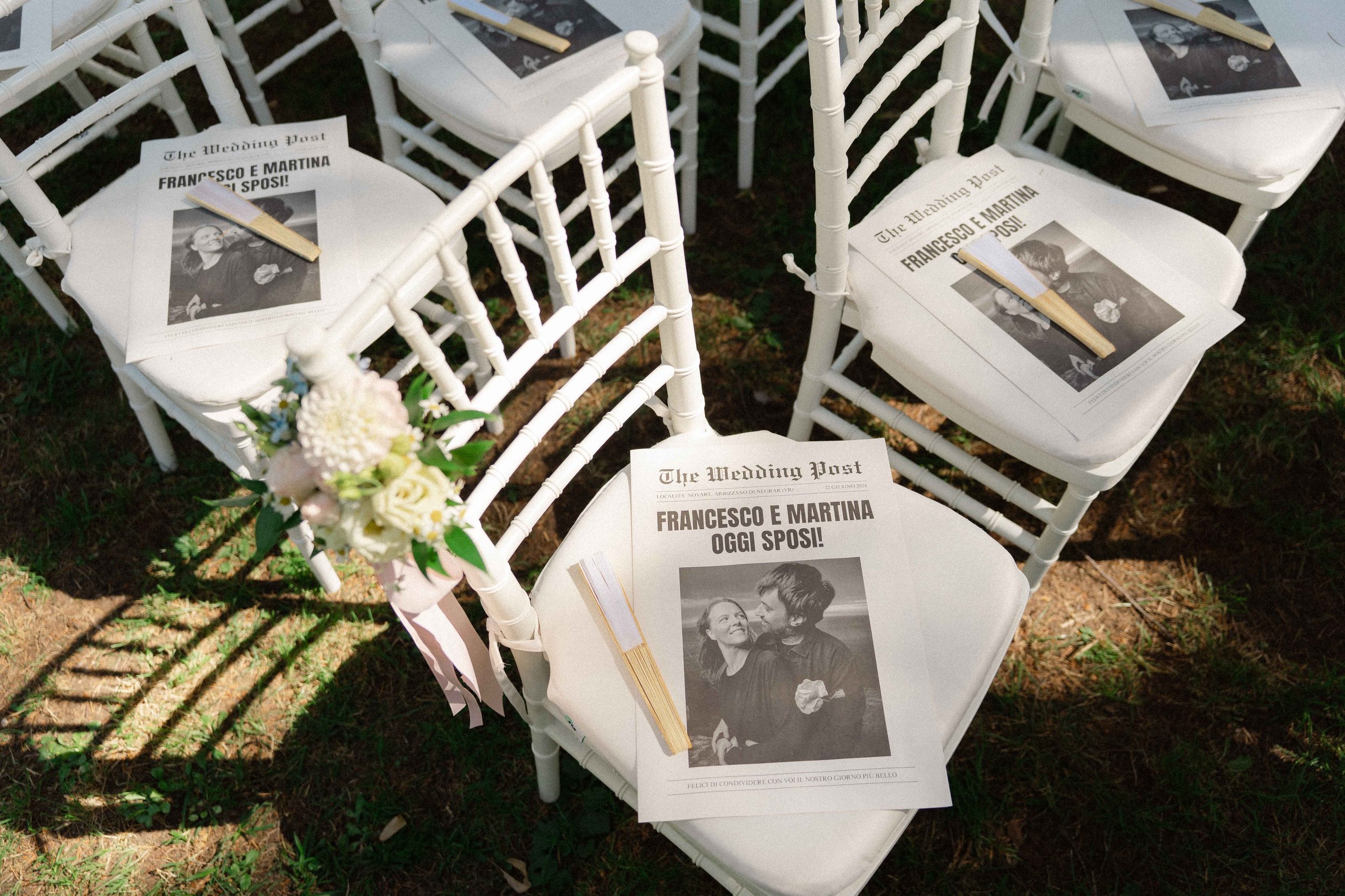 Wedding chairs decorated with printed programs on top, which feature a black and white photo of a couple and Italian text. One program includes a small fan. In the foreground, there is a bouquet of white and pink flowers.