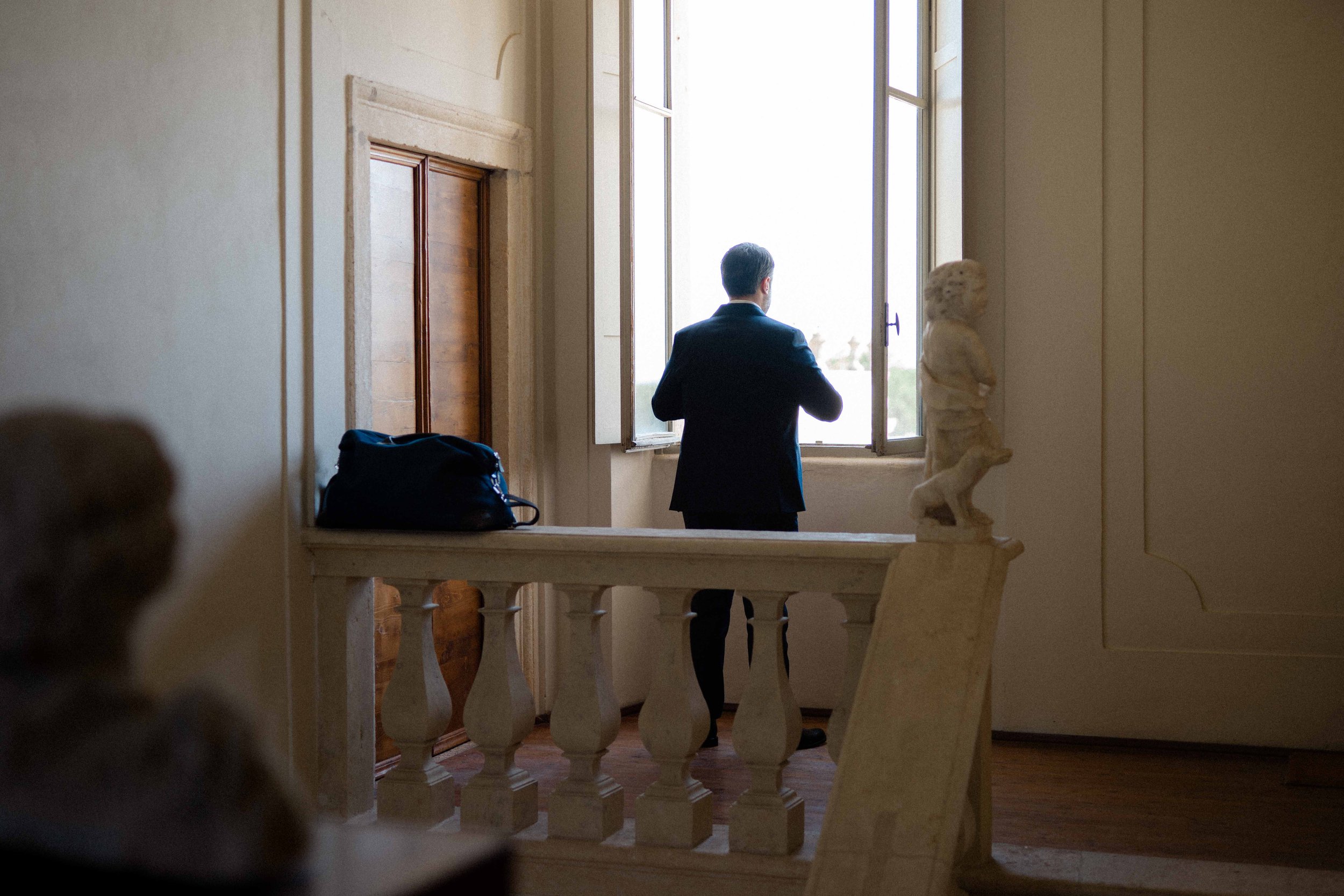 A man in a suit standing by an open window, looking outside. The room has a decorative bust on a pedestal and wooden flooring.