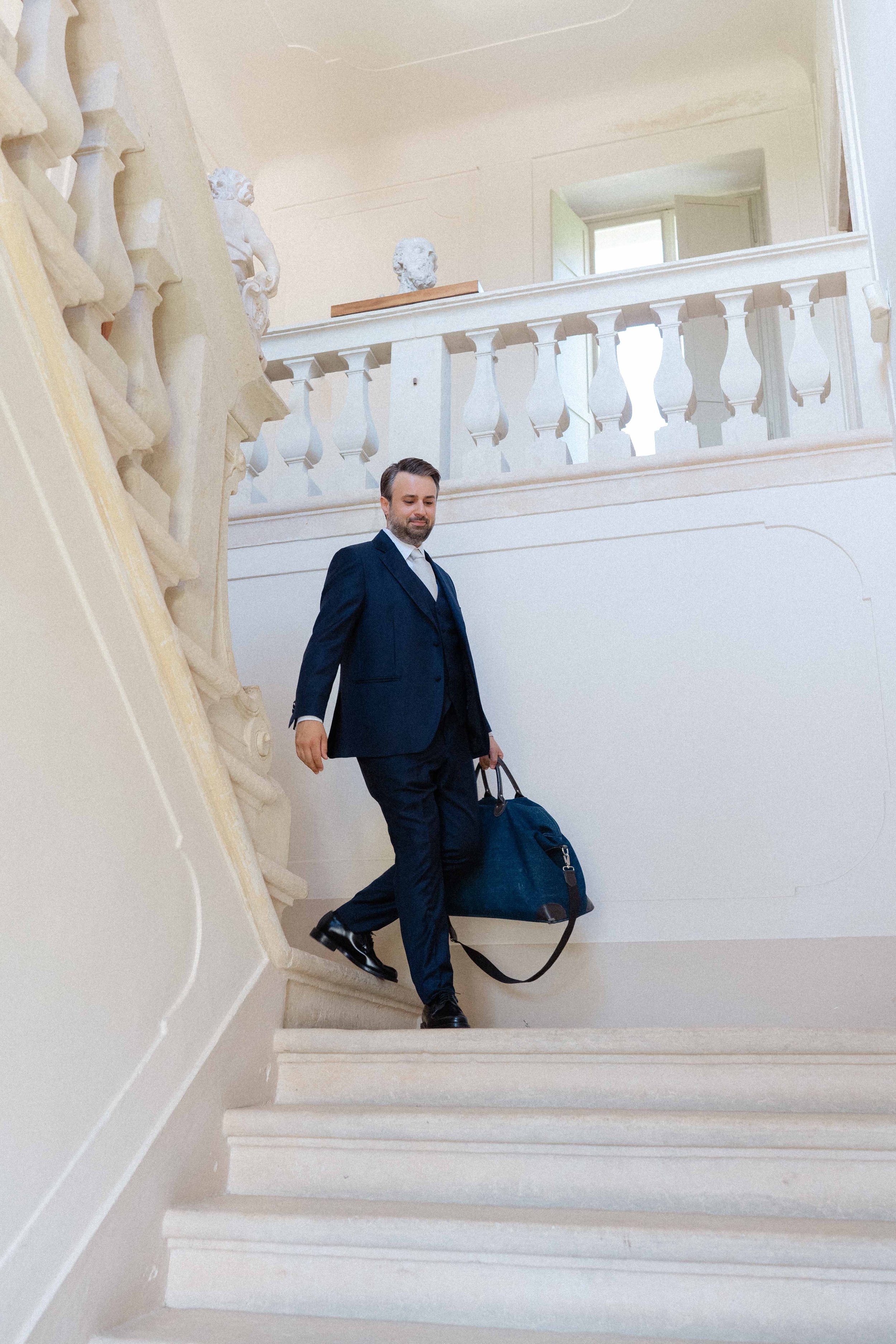 A man in a navy suit carrying a bag and walking down a staircase in a building with classical decor and sculptures.