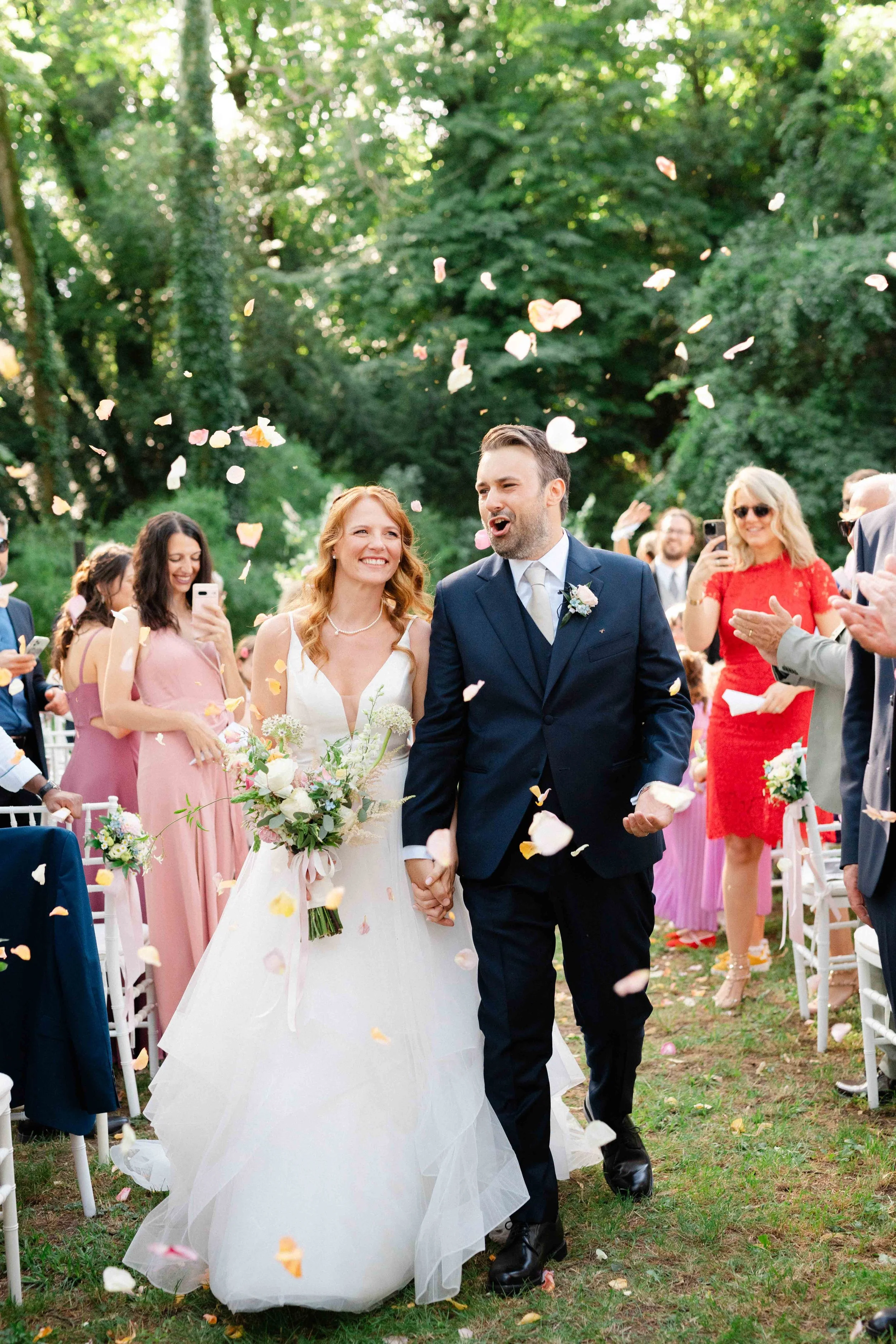 A bride and groom holding hands, walking down an outdoor aisle surrounded by friends and family. Festive flower petals are being tossed in the air as they smile and celebrate their wedding.